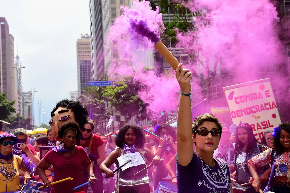 Imagem de manifestação nas ruas, uma mulher está a frente das demais segurando um sinalizador que solta fumaça roxa, há também um estandarte com a frase "Comida e Democracia", outras mulheres segurando bandeiras, instrumentos musicais feitos de lata de tinta e cabos de vassoura para fazer batucada. 
